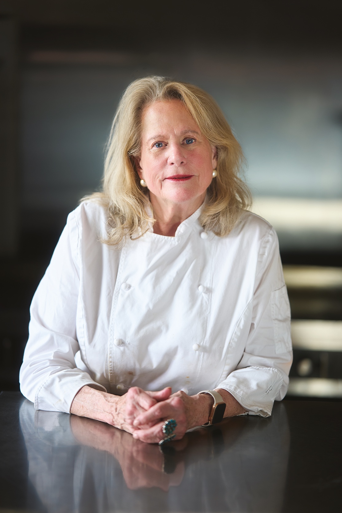 Nanette seated at a dark counter in chef whites, her hands folded, pearl earrings, looking straight at the reader with a gentle expression.