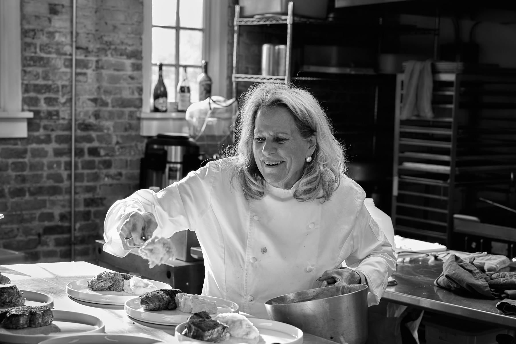 Black and white photograph: Nanette in her chef’s coat, smiling broadly, spoons potato onto a row of dinner plates in a brick-walled kitchen.