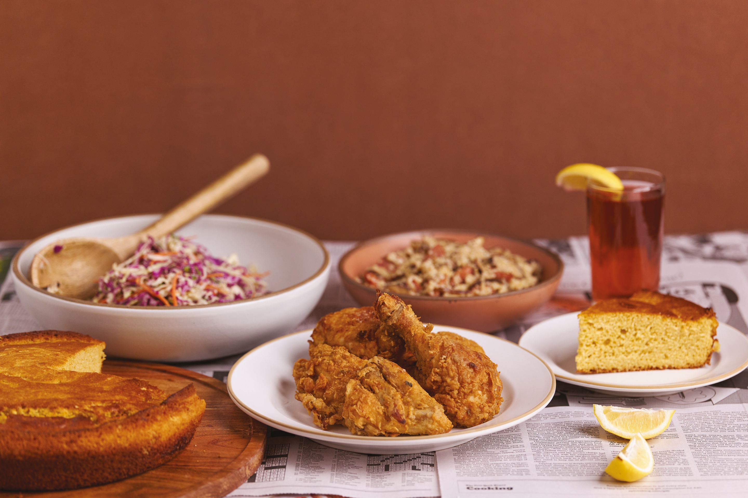 A supper laid on newspaper under a terracotta wall: a plate of fried chicken in front, bowls of coleslaw and grits behind it.