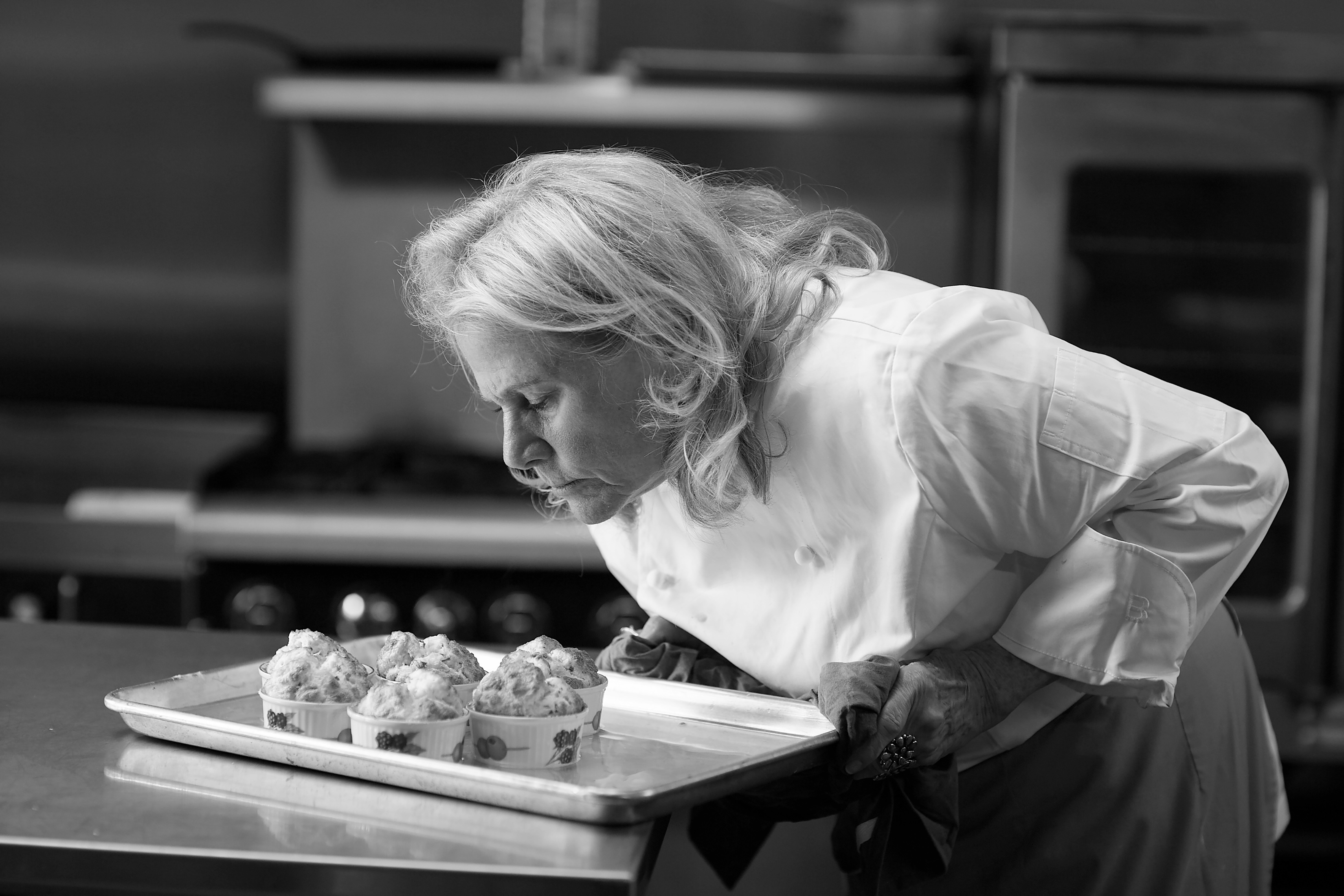 Black and white photograph: Chef Nanette Smalley bends low over a tray of muffins with her eyes closed, breathing them in.