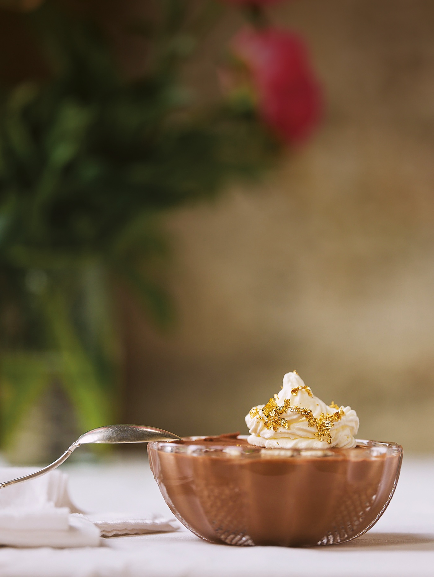 A cut-glass bowl of chocolate mousse topped with whipped cream and gold leaf, a silver spoon resting on its rim.