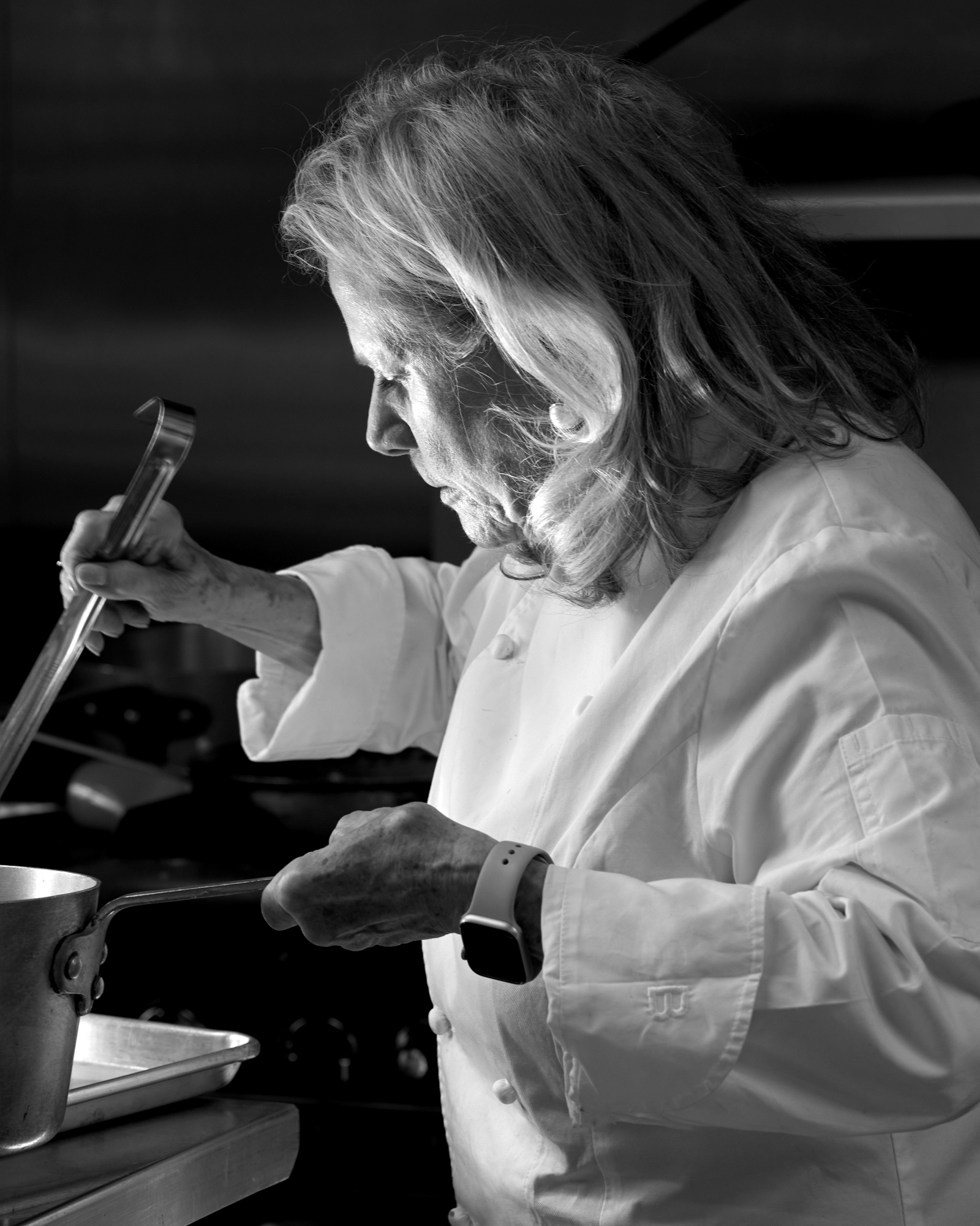 Black and white: Nanette in profile at the stove, head bowed, a long ladle raised in one hand and a saucepan’s handle in the other.