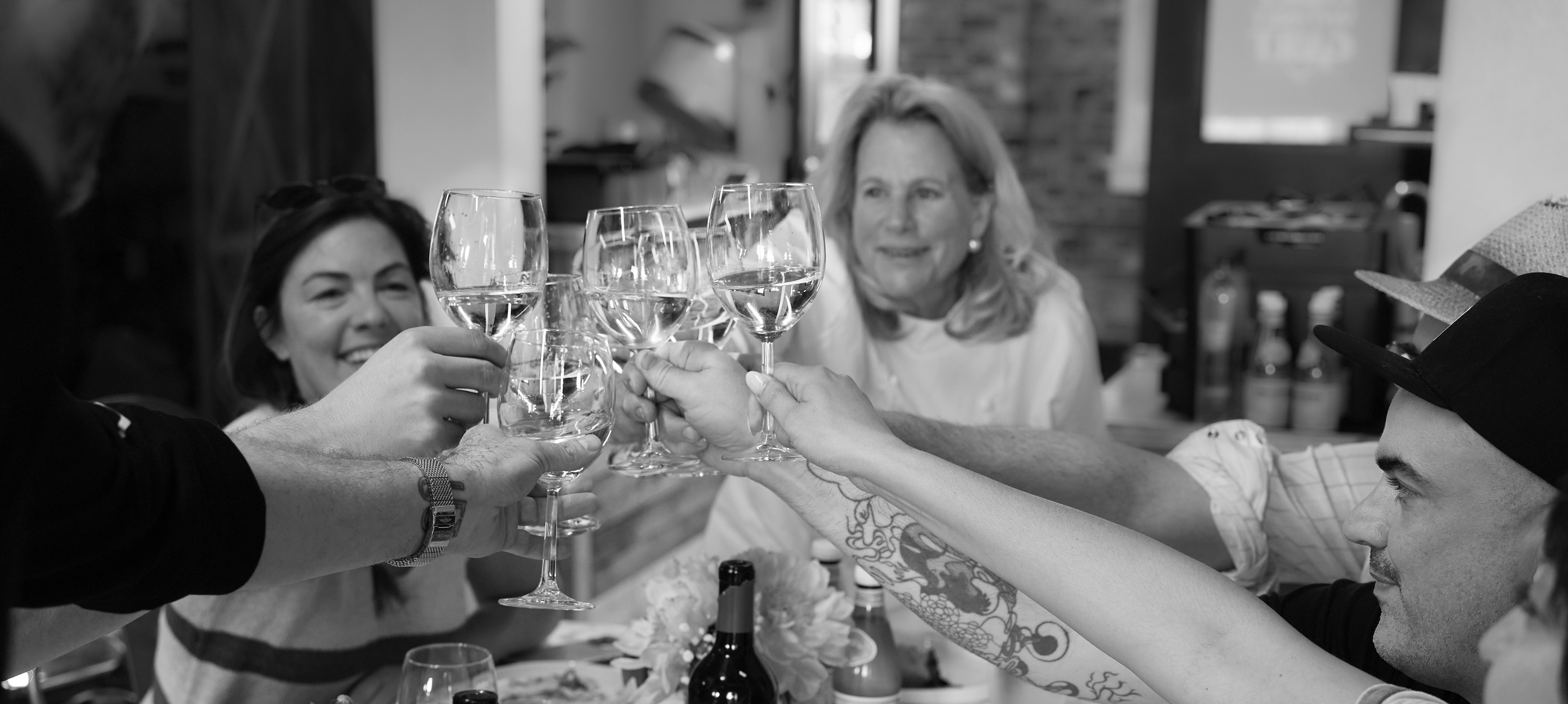 Black and white: wine glasses meet over a long table of plates and flowers; Nanette, at its head, smiles at the toast.
